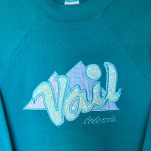 May include: Teal sweatshirt with a colorful embroidered design. The design features the word "Vail" in a light blue, textured font, with a mountain graphic and the word "Colorado" below. A vintage style.
