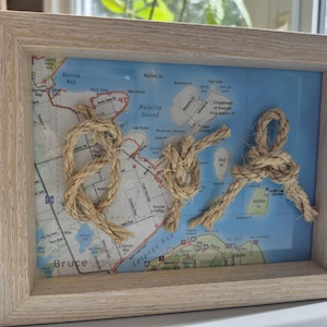 May include: A framed map art piece featuring a light-colored wooden frame. The map is of a coastal region, with three decorative rope knots attached to the map. The knots are made of natural-colored rope, adding a nautical theme.
