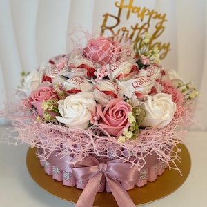 Könnte beinhalten: Ein festliches Geschenkarrangement mit einer Basis, die mit rosa Schleife und Schleife verziert ist. Es ist gefüllt mit verpackten Süßigkeiten, rosa und weißen Rosen und einem goldenen "Happy Birthday"-Topper. Das Farbschema ist rosa und weiß.