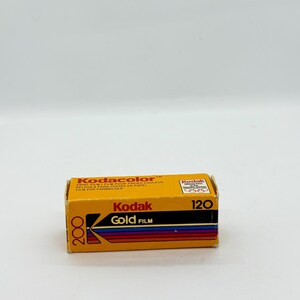 May include: A box of Kodak Gold 200 film. The box is yellow with black and red text, including "Kodak Gold Film" and "120". The film is designed for color prints. The box is rectangular and has a vintage aesthetic.