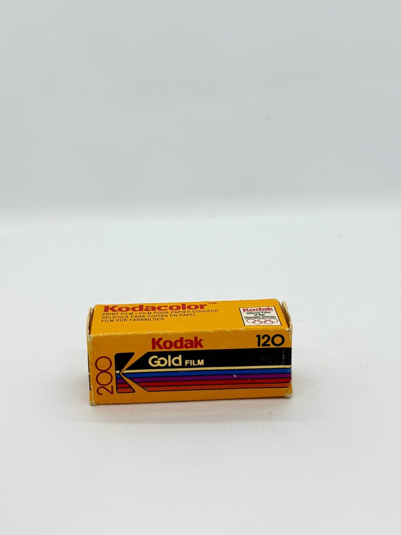 May include: A box of Kodak Gold 200 film. The box is yellow with black and red text, including "Kodak Gold Film" and "120". The film is designed for color prints. The box is rectangular and has a vintage aesthetic.