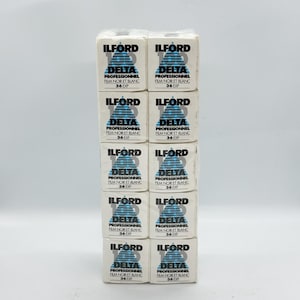 May include: A stack of eleven white film boxes. Each box features the brand name "ILFORD" in black, with "DELTA" in blue and white. The text "PROFESSIONNEL FILM NOIR ET BLANC 36 EXP" is also visible on each box.