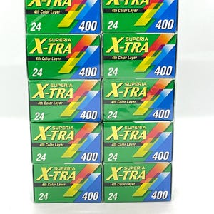 Puede incluir: Una pila de diez cajas de película Superia X-TRA 4th Color Layer. Cada caja verde presenta un diseño colorido y el texto "X-TRA", "24" y "400". Las cajas están dispuestas en tres filas.