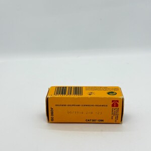 May include: A small, rectangular Kodak film box in yellow and black. The box has text that reads "ISO 200/24", "DEVELOP BEFORE", and "06/1994". The film box is a vintage item.