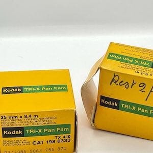 Puede incluir: Dos cajas amarillas de película Kodak TRI-X Pan. Las cajas tienen texto negro y detalles en verde. Una caja está abierta, revelando la película en su interior. Las cajas están etiquetadas con especificaciones de película, incluyendo 35 mm x 8,4 m.