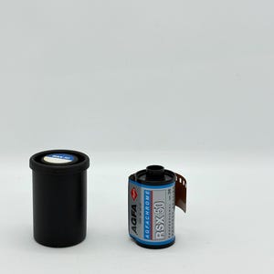 Può includere: Un contenitore nero per pellicola e un rotolo di pellicola AGFA RSX 50. Il rotolo di pellicola ha un'etichetta blu e argento con la scritta "AGFACHROME RSX 50". La pellicola è parzialmente srotolata, rivelando la striscia di pellicola.