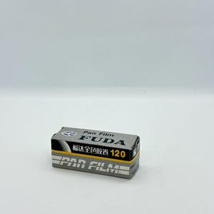 May include: A small, rectangular box of Fuda Pan Film 120, with a gray and black design. The box features the text "Pan Film" and "FUDA" in bold, white lettering, along with Chinese characters and the number "120".