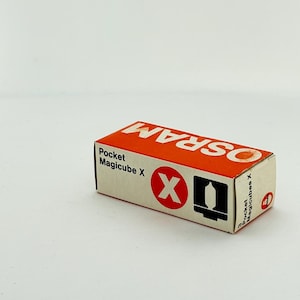以下が含まれることがあります： Osram Pocket Magicube Xフラッシュバルブ用の長方形の紙箱。パッケージは白地で、上部にはオレンジ色のパネルに白い文字でブランド名が記されています。前面には製品名、Xが描かれた赤い円、黒いアイコンが表示されています。