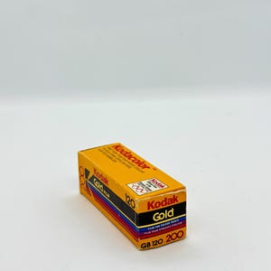 May include: A rectangular box of Kodak Gold 120 film. The box is primarily yellow with black, red, and blue accents. The Kodak Gold logo is prominently displayed, along with text indicating the film type and speed.