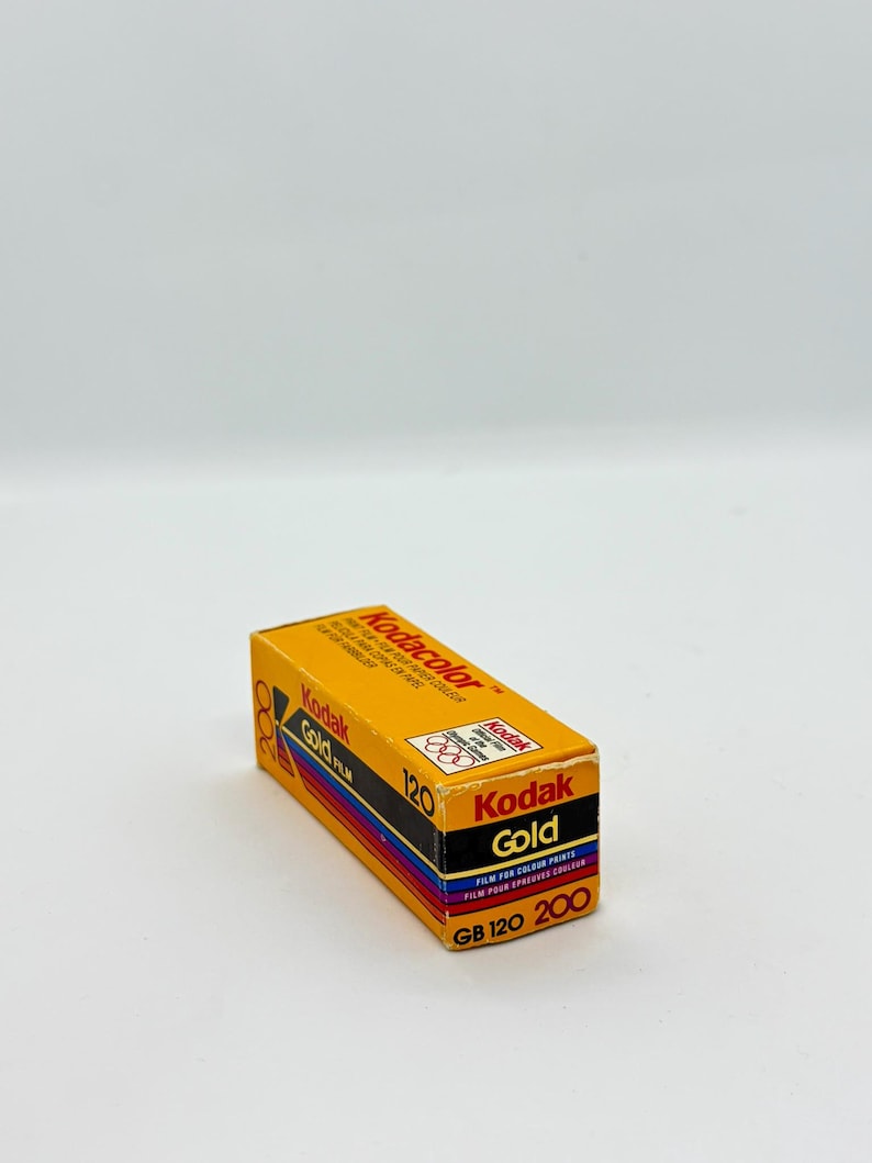 May include: A rectangular box of Kodak Gold 120 film. The box is primarily yellow with black, red, and blue accents. The Kodak Gold logo is prominently displayed, along with text indicating the film type and speed.