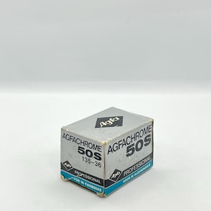 Peut inclure: Une petite boîte rectangulaire de film Agfachrome 50S. La boîte est principalement grise avec des accents noirs et turquoise. Le texte "AGFACHROME 50S" est bien en évidence, ainsi que "PROFESSIONAL" et "135-36". Le logo Agfa est visible.