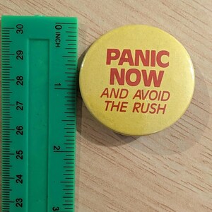 May include: A round yellow pin-back button with red text that reads PANIC NOW AND AVOID THE RUSH. The button is positioned next to a green plastic ruler showing a diameter of approximately 2.25 inches for the circular object.