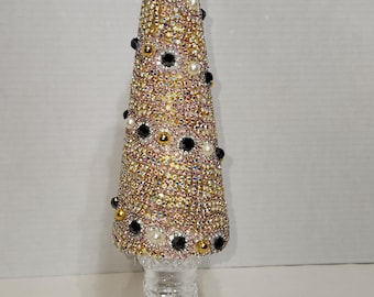 May include: A decorative, cone-shaped object covered in iridescent rhinestones, black and gold beads, and white pearls. The object sits atop a clear, cut-glass base, and is topped with a clear crystal. The overall design resembles a Christmas tree.