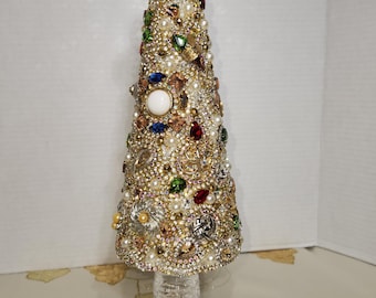 May include: A decorative Christmas tree made of gold-toned embellishments, pearls, and colorful jewels. The tree sits on a clear, cut-glass base and is topped with a faceted crystal. The tree is approximately 10 inches tall.
