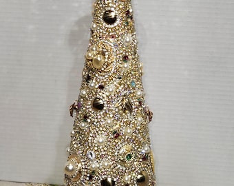 Puede incluir: Un árbol de Navidad decorativo, adornado con perlas, pedrería y detalles dorados. Está montado sobre una base de cristal tallado transparente y coronado por una esfera de cristal. Los colores dominantes son el dorado y el blanco.