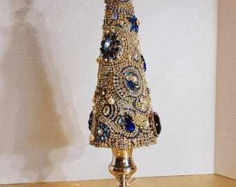 May include: A decorative Christmas tree made of gold and silver rhinestones, blue jewels, and pearls. The tree sits on a silver candlestick base. The tree is approximately 12 inches tall.