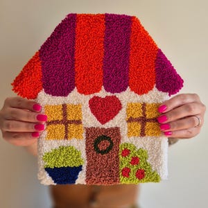 Pode incluir: Um pendurador de parede em forma de casa, colorido e texturizado. O telhado apresenta listras laranja e roxas, com um corpo branco. Detalhes: janelas amarelas, coração vermelho, porta marrom e arbusto verde com bagas vermelhas. Um vaso azul com plantas verdes também é visível.