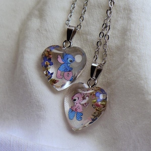 May include: Two heart-shaped pendants with silver chains. Each pendant features a cartoon character inside a clear resin heart, surrounded by small flowers. One pendant has a blue character, the other a pink one.