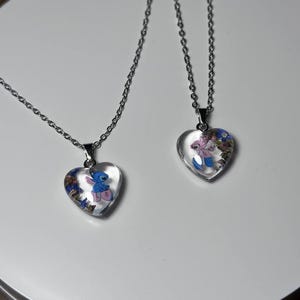 Puede incluir: Dos collares con colgantes en forma de coraz&oacute;n y cadenas plateadas. Cada colgante contiene un personaje de dibujos animados y detalles florales coloridos. Los colgantes son transparentes, lo que permite ver el dise&ntilde;o. Los collares se exhiben sobre una superficie blanca.