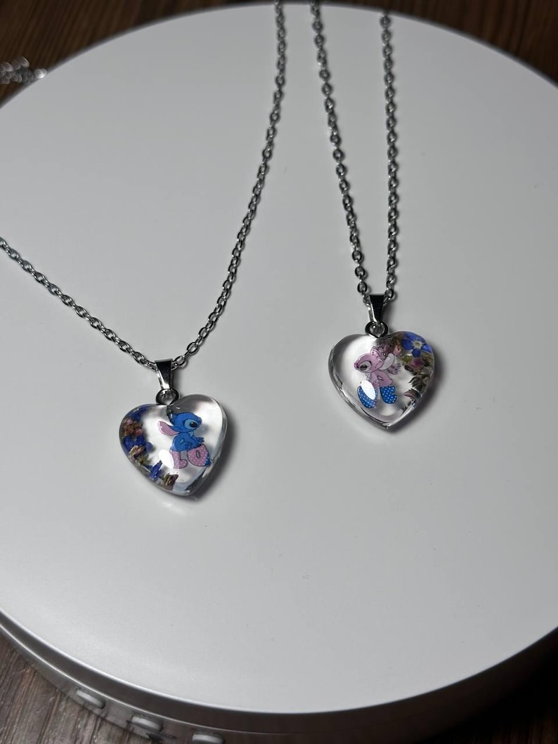 Puede incluir: Dos collares con colgantes en forma de coraz&oacute;n y cadenas plateadas. Cada colgante contiene un personaje de dibujos animados y detalles florales coloridos. Los colgantes son transparentes, lo que permite ver el dise&ntilde;o. Los collares se exhiben sobre una superficie blanca.