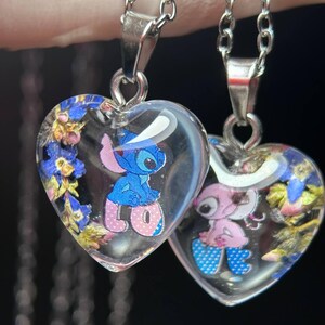 Puede incluir: Dos colgantes en forma de coraz&oacute;n con resina transparente, cada uno con un personaje de dibujos animados y flores secas. Los colgantes cuelgan de cadenas plateadas. Un colgante tiene un personaje azul, el otro rosa.