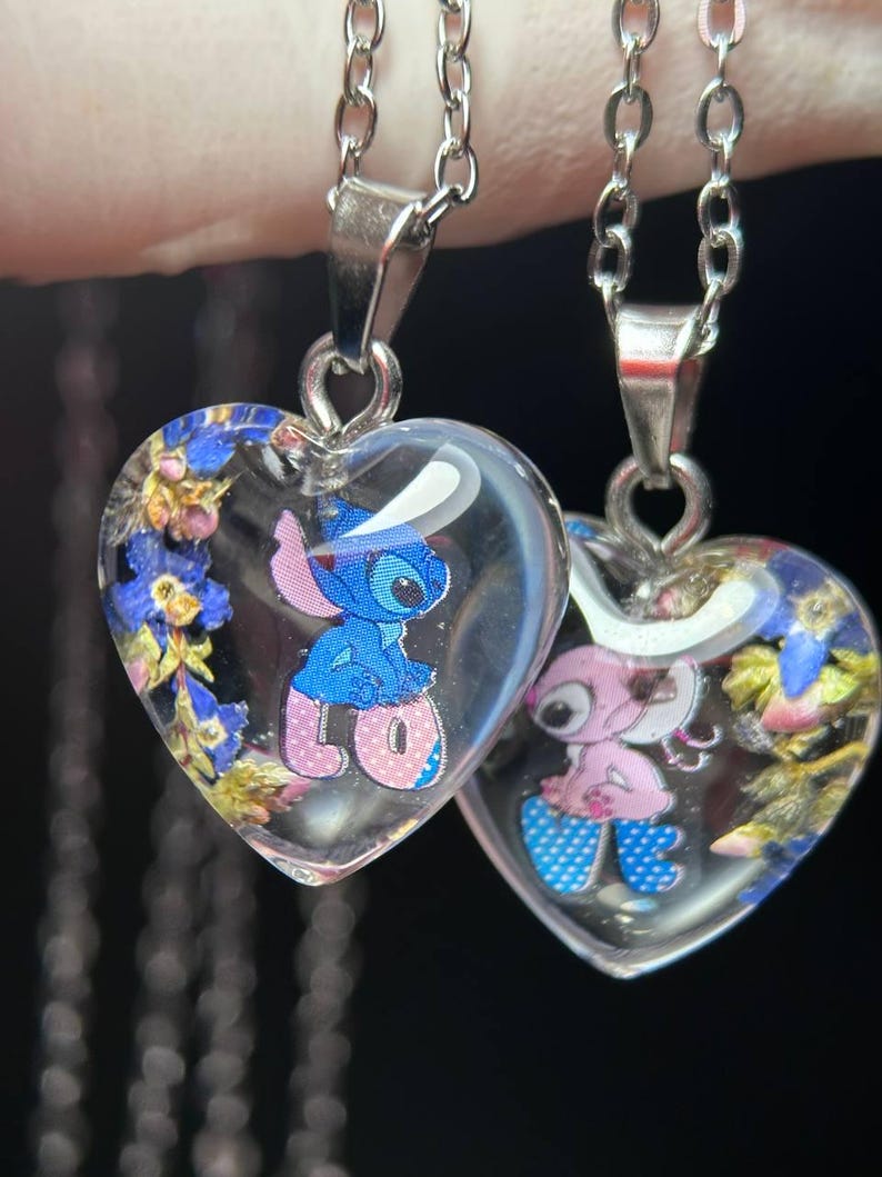 Puede incluir: Dos colgantes en forma de coraz&oacute;n con resina transparente, cada uno con un personaje de dibujos animados y flores secas. Los colgantes cuelgan de cadenas plateadas. Un colgante tiene un personaje azul, el otro rosa.