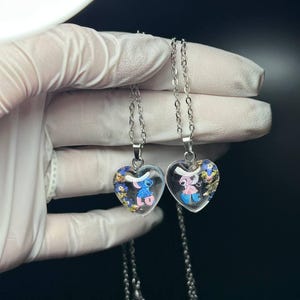 Puede incluir: Dos colgantes en forma de coraz&oacute;n con cadenas plateadas. Cada colgante contiene un personaje de dibujos animados, uno azul y otro rosa, rodeado de detalles coloridos. Los colgantes son transparentes.