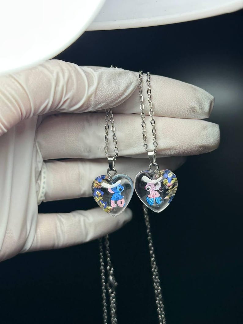 Puede incluir: Dos colgantes en forma de coraz&oacute;n con cadenas plateadas. Cada colgante contiene un personaje de dibujos animados, uno azul y otro rosa, rodeado de detalles coloridos. Los colgantes son transparentes.