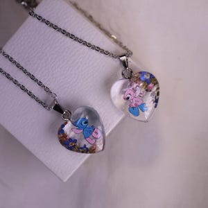 Puede incluir: Dos colgantes en forma de coraz&oacute;n con cadenas de plata. Cada colgante presenta un dise&ntilde;o de personaje de dibujos animados con acentos florales coloridos encerrados en resina transparente. Los colgantes se exhiben sobre una superficie blanca.