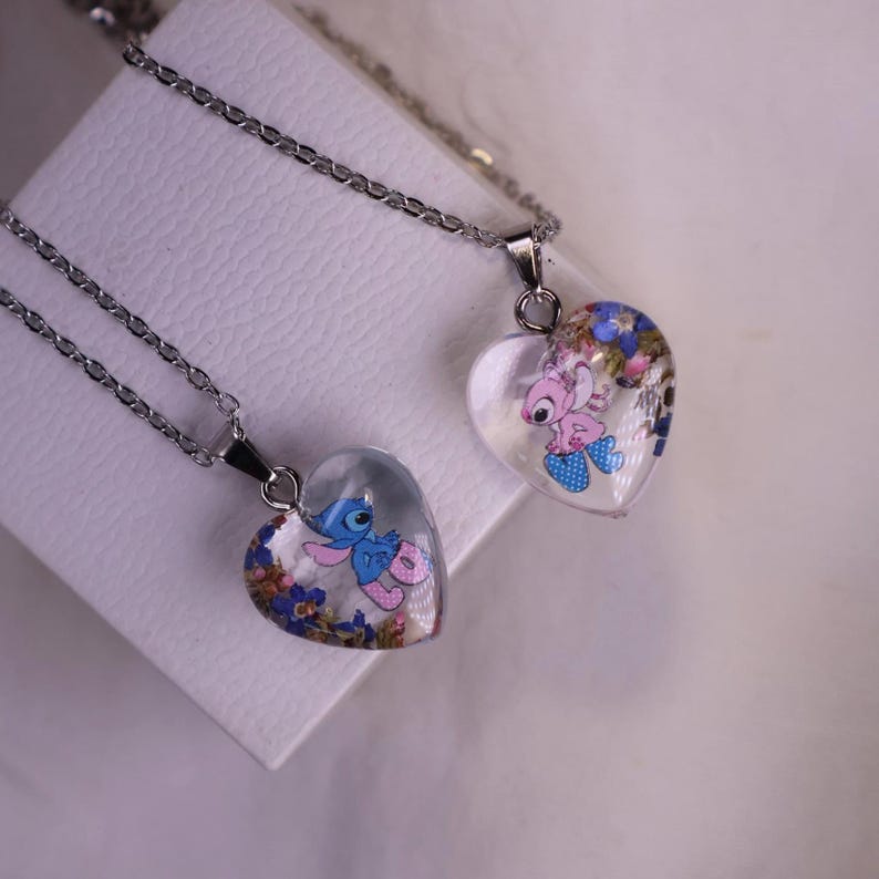 Puede incluir: Dos colgantes en forma de coraz&oacute;n con cadenas de plata. Cada colgante presenta un dise&ntilde;o de personaje de dibujos animados con acentos florales coloridos encerrados en resina transparente. Los colgantes se exhiben sobre una superficie blanca.