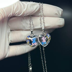 Puede incluir: Dos colgantes en forma de coraz&oacute;n con cadenas plateadas. Cada colgante contiene un personaje de dibujos animados y peque&ntilde;as flores azules y amarillas. Un colgante presenta un personaje azul, el otro un personaje rosa.