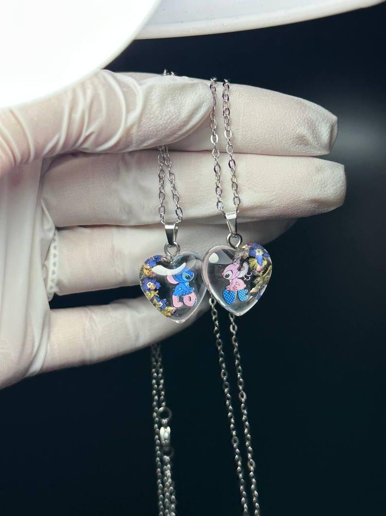 Puede incluir: Dos colgantes en forma de coraz&oacute;n con cadenas plateadas. Cada colgante contiene un personaje de dibujos animados y peque&ntilde;as flores azules y amarillas. Un colgante presenta un personaje azul, el otro un personaje rosa.