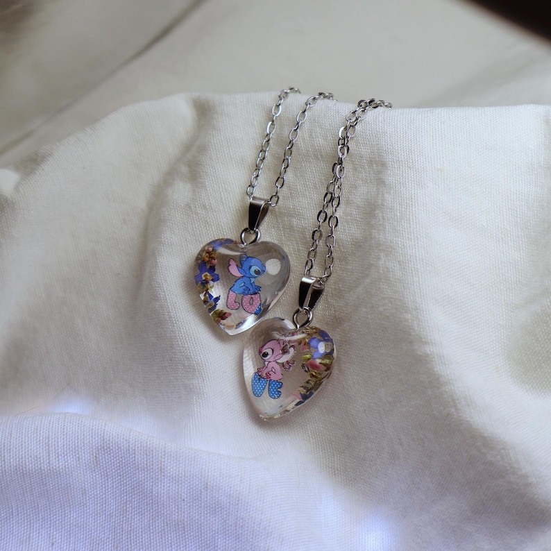 May include: Two heart-shaped pendants with silver chains. Each pendant features a cartoon character inside a clear resin heart, surrounded by small flowers. One pendant has a blue character, the other a pink one.