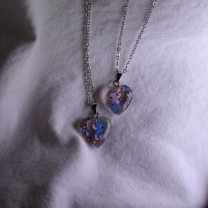 Puede incluir: Dos colgantes en forma de coraz&oacute;n con personajes de dibujos animados en cadenas de plata. Los colgantes transparentes contienen confeti de colores y presentan un personaje de dibujos animados azul y rosa. Sobre un fondo de tela blanca.