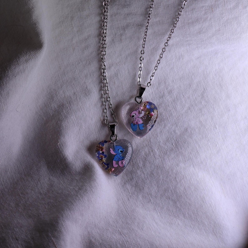 Puede incluir: Dos colgantes en forma de coraz&oacute;n con personajes de dibujos animados en cadenas de plata. Los colgantes transparentes contienen confeti de colores y presentan un personaje de dibujos animados azul y rosa. Sobre un fondo de tela blanca.