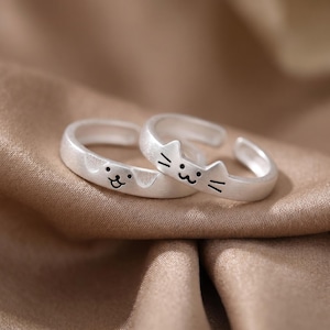 Könnte beinhalten: Zwei silberfarbene offene Ringe mit matter Oberfläche. Ein Ring zeigt ein eingraviertes Hundegesicht mit Schlappohren, während der andere Ring ein eingraviertes Katzengesicht mit spitzen Ohren und Schnurrhaaren aufweist.