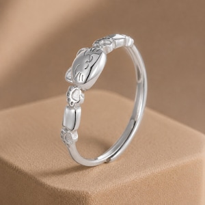 May include: A silver-toned ring featuring a polished cat face design at the center, flanked by small engraved cat paw prints and smooth rectangular links along the band. The metal has a reflective finish and a continuous, open-ended circular shape.