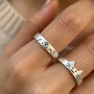 May include: Two silver rings, one with a cat face design and the other with a cat silhouette. The rings are worn on fingers, showcasing the minimalist and cute cat-themed jewelry.