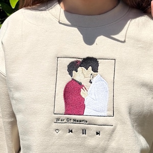 May include: Beige sweatshirt with an embroidered design featuring two figures kissing, with the text "War Of Hearts" below. The design is framed within a square, resembling a music player interface. The person in the image is wearing sunglasses.