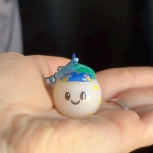 May include: A small, round, white keychain with a smiling face and a blue and green hat. The keychain has a small loop at the top for attaching to keys or a bag. The design includes yellow and blue accents.