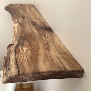 May include: A live edge wooden wall shelf featuring a natural light brown grain with dark brown knots and organic wood patterns. The shelf has a smooth, finished surface and an irregular, raw edge that follows the original shape of the wood.