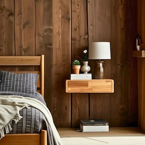 May include: A wooden bedside table with a drawer, a white lamp with a white shade, a small potted plant, and a stack of books. The table is mounted to a wood wall.