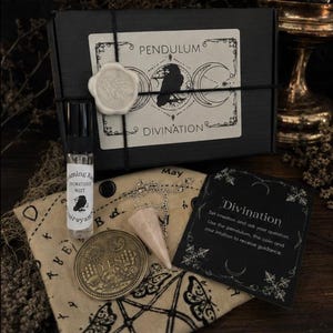 Könnte beinhalten: Eine schwarze Schachtel mit der Aufschrift Pendulum Divination, einem weißen Wachssiegel, einer kleinen Sprühflasche, einer runden Metallmünze mit dem Wort YES, einem hellrosa Kristallpendel an einer Silberkette und einer Anleitung auf beigem Stoff.