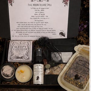 Könnte beinhalten: Eine schwarze Geschenkbox mit einer Anleitung für einen Full Moon Release Spell, einem Sleepy Lavender-Etikett, einer Sprühflasche mit Luna Mist-Aufschrift, einem schwarzen Beutel mit Steinen, einer Kerze in einer Dose und Kräutern.