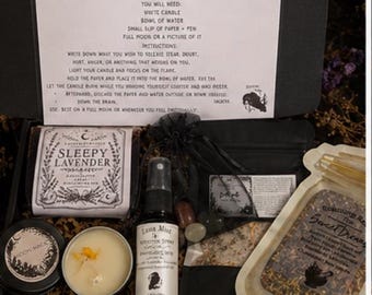 Op de afbeelding: Een zwarte geschenkdoos met een instructiekaart voor een Full Moon Release Spell, een Sleepy Lavender-label, een sprayflesje met het label Luna Mist, een zwart zakje met stenen, een kaars in een blikje en een zakje met gedroogde kruiden.