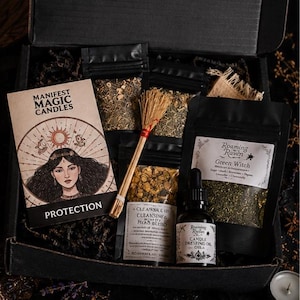 May include: A black box containing a Manifest Magic Candles Protection card, a small straw broom, three black pouches of dried herbs, and a glass dropper bottle labelled Candle Dressing Oil. The items are arranged inside the box with dark filler.