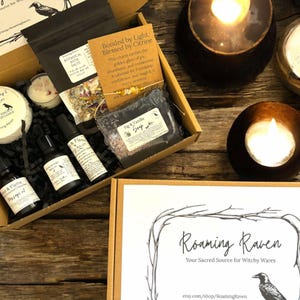 May include: A brown box with a black and white raven logo and the text "Roaming Raven Your Sacred Source for Witchy Wares etsy.com/shop/RoamingRaven". The box contains a variety of witchy items, including bath salts, a soap bar, a candle, and essential oils.