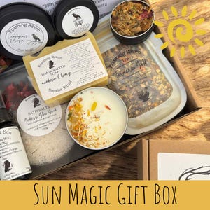 May include: A Sun Magic gift box filled with self-care items. Includes soap, bath salts, tea, a candle, and other products. The box has a wooden background and a yellow sun graphic.