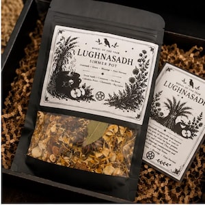 May include: A black resealable pouch containing dried botanicals labelled Lughnasadh Simmer Pot with a white paper label featuring black ink illustrations of a cauldron, wheat, and text listing ingredients like dried apple and cinnamon.