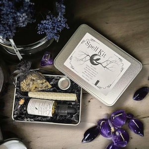Puede incluir: Una caja de metal plateada con las palabras "Spell Kit" impresas en la tapa. La caja contiene una vela pequeña, una botella de aceite, un cristal y una bolsa pequeña. La caja está sobre una mesa de madera.